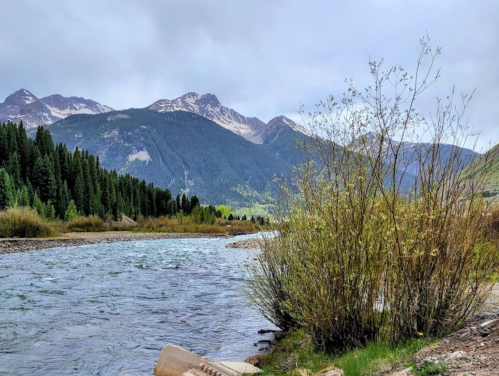 A serene river flows through a valley surrounded by mountains and lush greenery under a cloudy sky.