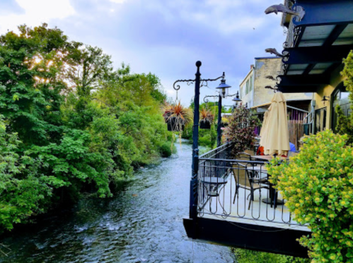 A scenic view of a river beside a patio with greenery and trees, under a partly cloudy sky.
