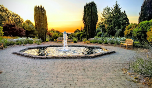 A serene garden at sunset featuring a fountain, benches, and lush greenery.