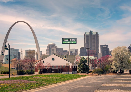 A view of a city skyline with a large arch, trees, and a billboard in the foreground on a clear day.