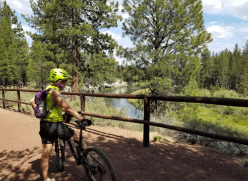 A person in a bright helmet stands beside a mountain bike, overlooking a river surrounded by trees.