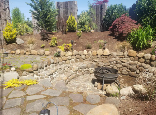 A serene garden with stone pathways, a fire pit, and lush greenery, surrounded by trees and colorful plants.