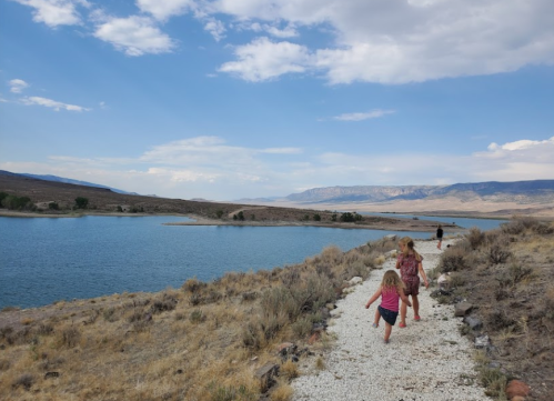 Two children run along a gravel path by a lake, with mountains and a blue sky in the background.