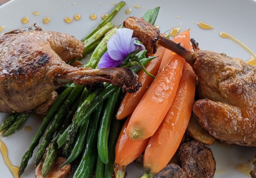 A plated dish featuring roasted quail, green beans, carrots, and garnished with a purple flower.