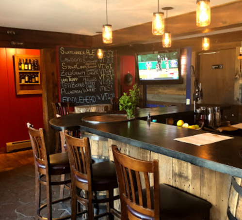 A cozy bar with wooden stools, a chalkboard menu, and a TV, featuring warm lighting and rustic decor.