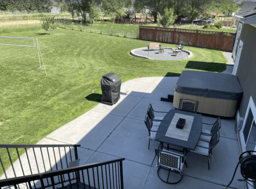 A spacious backyard with a patio, hot tub, grill, and a fire pit area surrounded by grass and trees.