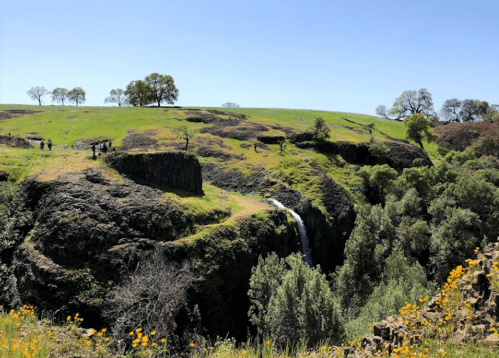 A scenic landscape featuring a waterfall cascading down rocky cliffs, surrounded by green hills and blooming wildflowers.