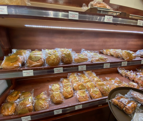 A display of various baked goods in clear packaging on wooden shelves, labeled with prices.