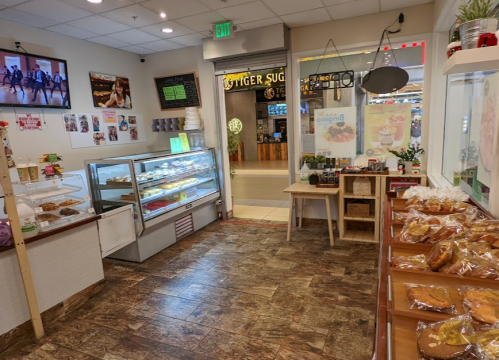 A cozy shop interior with a display case of pastries, a snack counter, and a screen showing a dance performance.