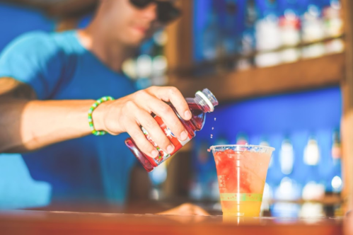 A person in a blue shirt pours a colorful drink into a cup at a bar, with shelves of bottles in the background.