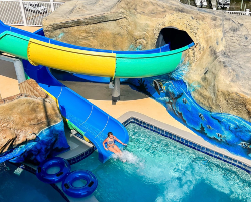 A child slides down a colorful water slide into a pool surrounded by a rock formation and blue water.
