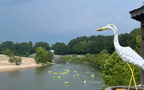 A white heron statue overlooks a river with kayakers and lush green trees under a cloudy sky.