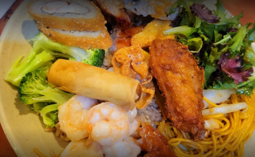 A colorful plate of food featuring shrimp, chicken wings, spring rolls, broccoli, and mixed greens over rice and noodles.