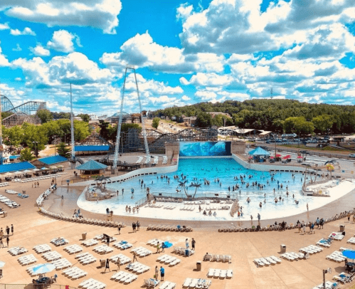 A vibrant water park scene with a large pool, people swimming, and amusement rides under a blue sky with fluffy clouds.