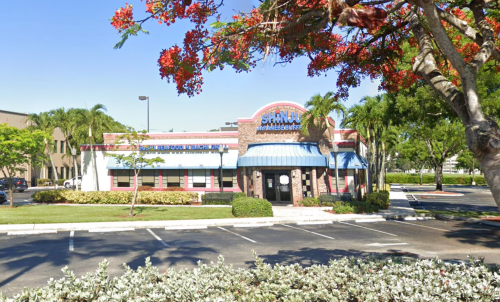 A restaurant with a blue awning and red brick exterior, surrounded by palm trees and colorful flowers.