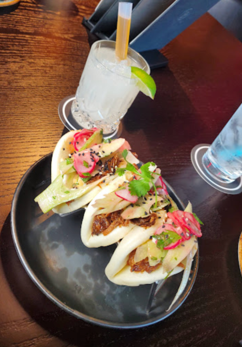 Three bao buns filled with savory toppings, accompanied by a cocktail with a lime wedge on a wooden table.