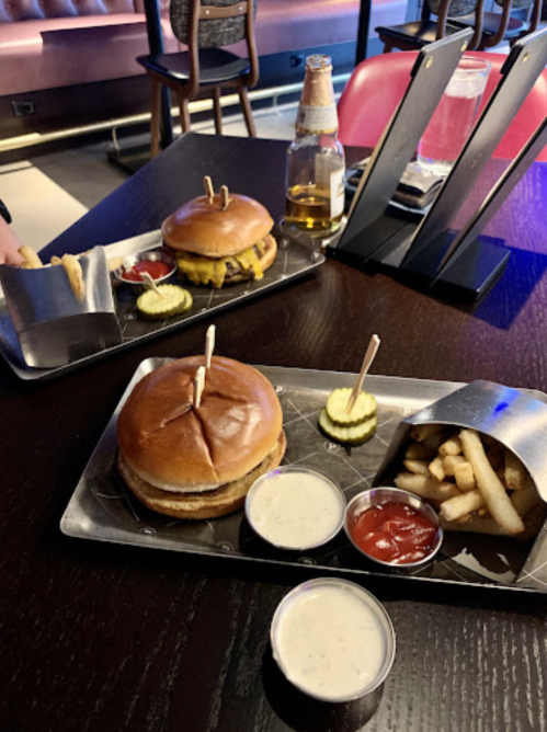 Two trays with burgers, pickles, fries, and dipping sauces on a dark wooden table in a casual dining setting.