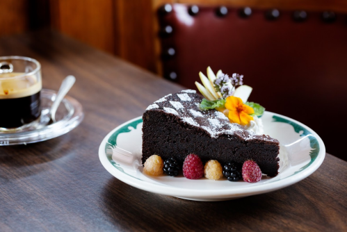 A slice of chocolate cake garnished with fruit and flowers, served on a plate next to a cup of coffee.