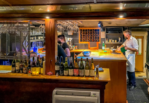 A cozy restaurant kitchen with staff preparing food, shelves of wine bottles, and a warm, inviting atmosphere.