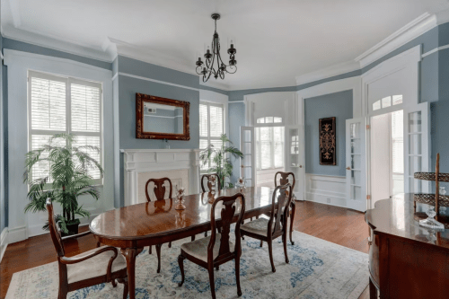 Elegant dining room with a wooden table, chandelier, and large windows, featuring blue walls and decorative plants.