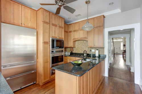 Modern kitchen with wooden cabinets, stainless steel appliances, and a dark countertop, leading to a hallway.