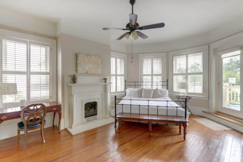 Bright bedroom with a bed, wooden floors, a fireplace, and large windows. A desk is in the corner.
