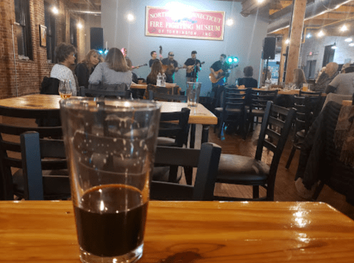 A glass of dark beer sits on a wooden table, with a live band performing in the background at a lively venue.