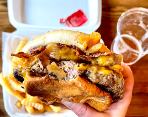 A hand holding a loaded cheeseburger with bacon, melted cheese, and onions, alongside crinkle-cut fries and a drink.