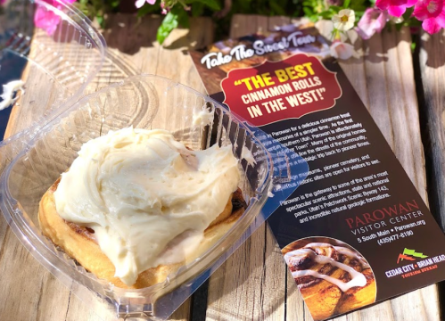 A cinnamon roll with cream cheese frosting in a takeout container, next to a promotional flyer for a visitor center.