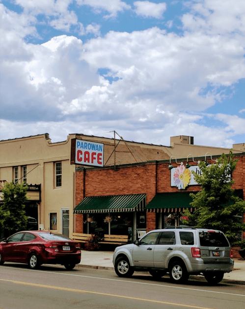 A street view of Parowan Cafe with a blue sky and clouds, surrounded by trees and parked cars.