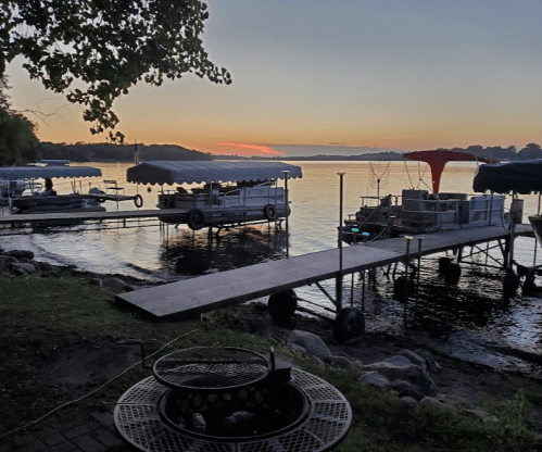 A serene lakeside scene at sunset, featuring docks, boats, and a fire pit on the shore.