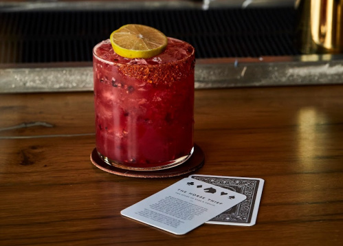 A vibrant cocktail with a lime slice on top, next to a playing card on a wooden table.