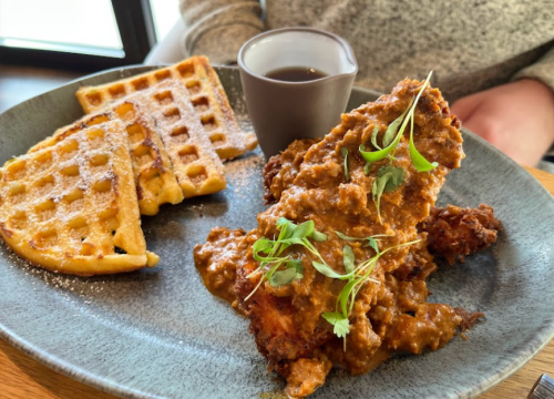 A plate featuring fried chicken topped with gravy, served with waffles and a small cup of syrup.