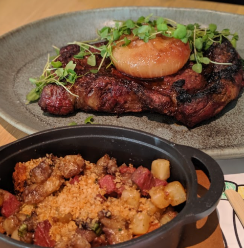 A grilled steak topped with a caramelized onion and microgreens, served with a side of savory stuffing in a small dish.