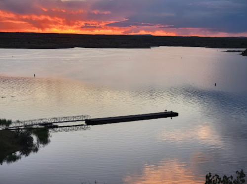 A serene lake at sunset, reflecting vibrant orange and purple hues, with a small dock extending into the water.