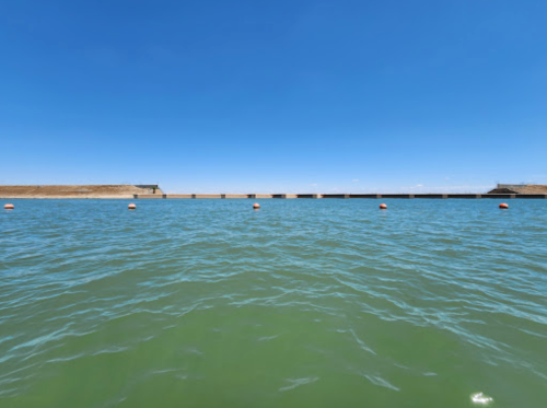 A calm body of water with a clear blue sky and a distant shoreline, marked by floating buoys.