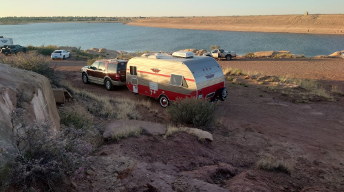 A vintage camper parked near a lake, surrounded by rocky terrain and sparse vegetation.