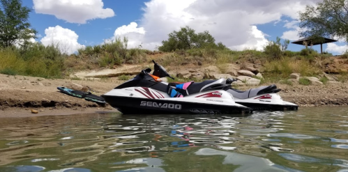 Two jet skis parked on the shore of a calm lake, with trees and clouds in the background.
