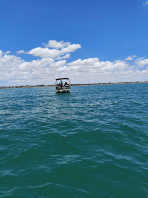 A small boat with two people is seen on a calm blue lake under a bright sky with fluffy clouds.
