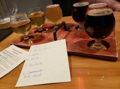 A wooden flight of six craft beer samples with a handwritten note listing their names on a table.