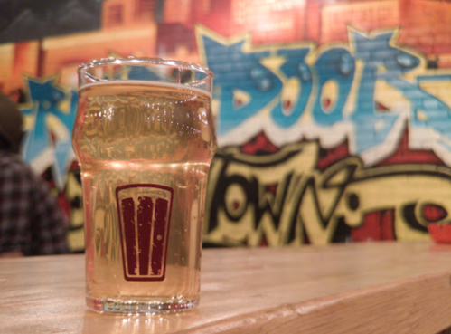 A glass of beer sits on a wooden table in front of a colorful graffiti wall.