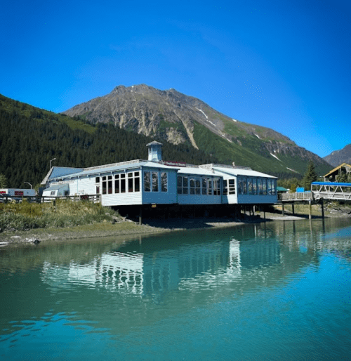 A waterfront building with large windows reflects in calm water, surrounded by mountains and clear blue skies.