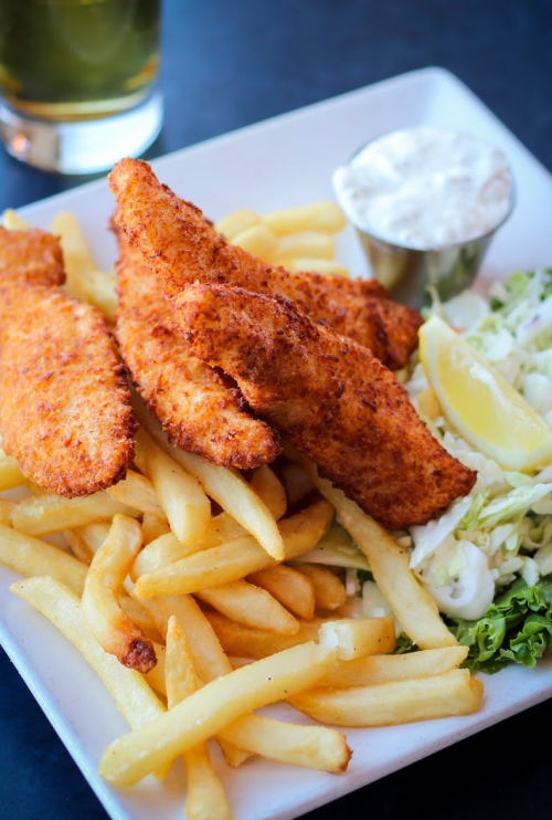 A plate of crispy fried fish fillets served with French fries, coleslaw, a lemon wedge, and a small cup of dipping sauce.