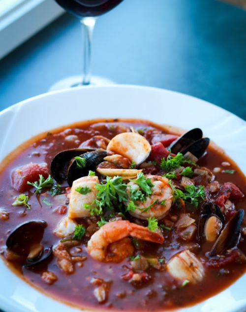 A bowl of seafood stew with shrimp, mussels, and herbs, served with a glass of red wine in the background.