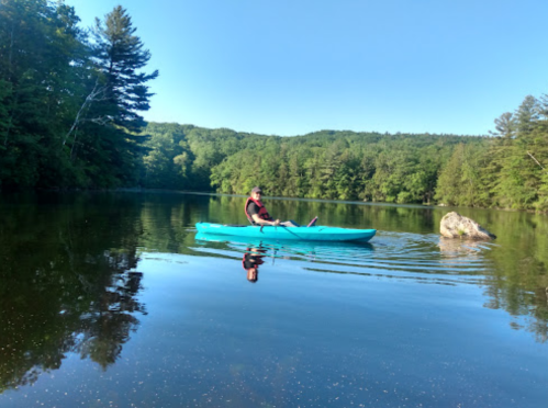 A person in a turquoise kayak paddles on a calm lake surrounded by lush green trees and a rock.
