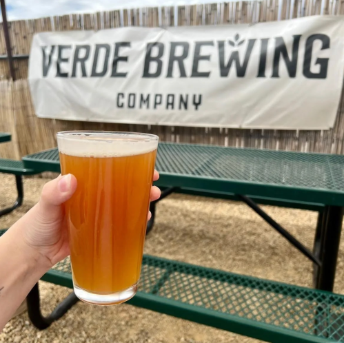 A hand holds a pint of amber beer in front of a Verde Brewing Company banner and picnic tables.