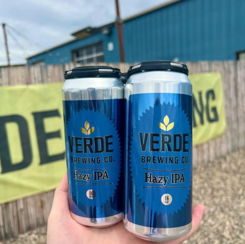 Two cans of Verde Brewing Co. Hazy IPA held in hand, with a blue background and a brewery sign in the distance.
