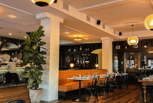 A modern restaurant interior featuring a cozy booth, bar area, and decorative plants under warm lighting.