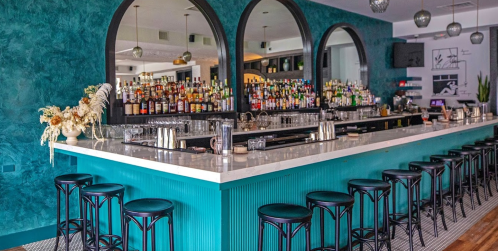 A stylish bar with a turquoise counter, black stools, and a well-stocked shelf of various liquor bottles in the background.