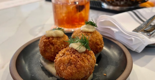 Three golden-brown croquettes topped with dill and sauce, served on a plate with a cocktail in the background.